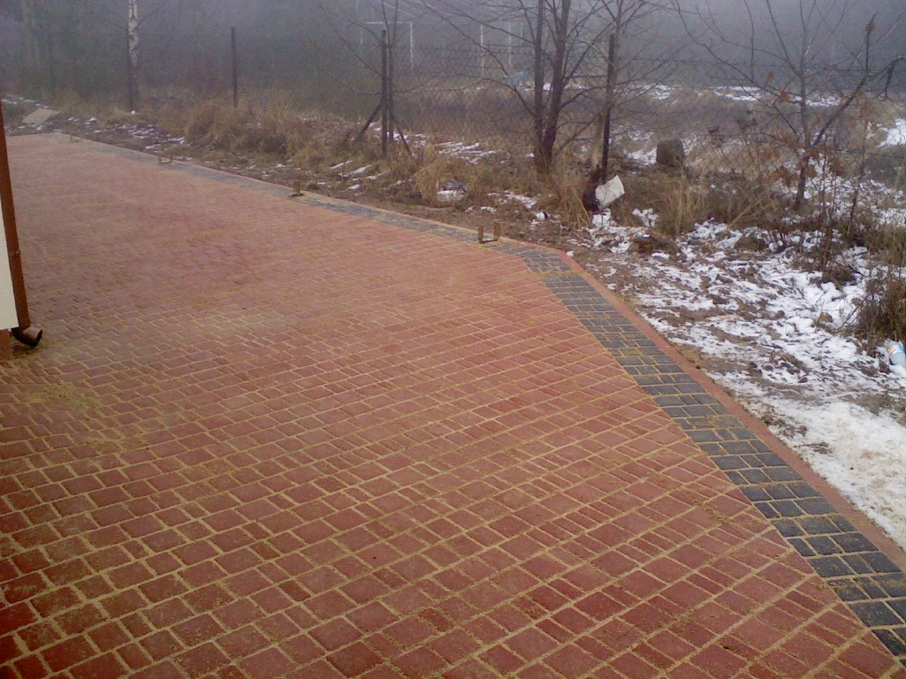 Nowo ułożona, czerwona kostka brukowa w jodełkę z ciemnoszarym obramowaniem, widoczna w pochmurny dzień na tle ogrodu z resztkami śniegu.